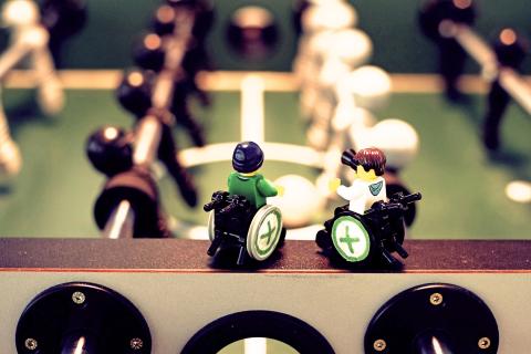 Zwei im Rollstuhl sitzende Legofiguren stehen auf dem Rand eines Kickertisches und schauen auf das Spielfeld mit schwarzen und weißen Spielerfiguren