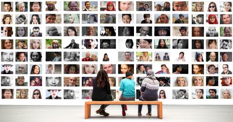 Drei Menschen sitzen in einer Galerie auf einer Holzbank und schauen auf eine Wand voller Fotos mit Aufnahmen verschiedener Gesichter