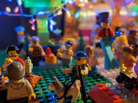 Szene aus Lego-Figuren, die ein buntes Fest darstellt, auf der Menschen unterschiedlichsten Alters und Geschlechts, mit ausgefallenen Kostümen, Assistenzhunden etc. sind