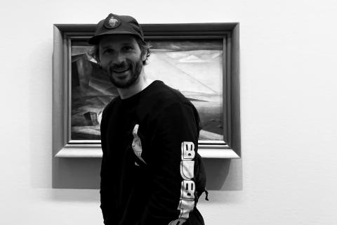 Mann mit Bart, lässigem Longsleeve und Baseball-Mütze steht vor einem abstrakten Gemälde und lacht in die Kamera