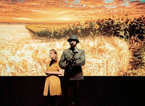 Bühnenbild eines Weizenfeldes mit Sonnenblumen, davor eine Frau im Blumenkleid und ein Soldat auf der Bühne